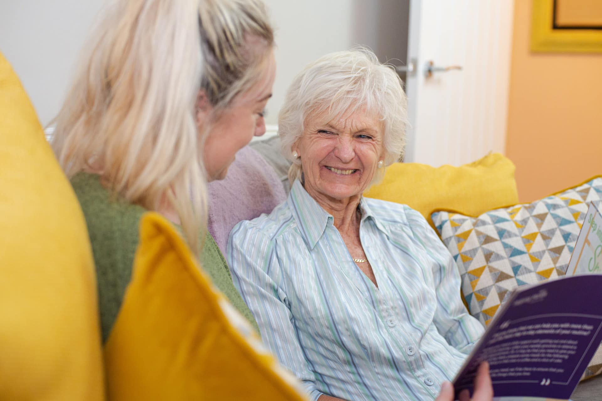 Elderly lady smiling shirt female carer green top yellow cushions